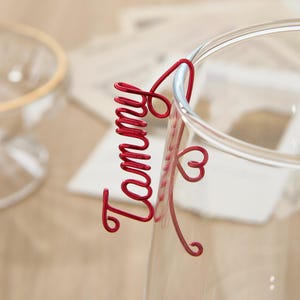May include: A red wire name tag with the word "Tammy" and a heart shape. The tag is attached to a clear glass.