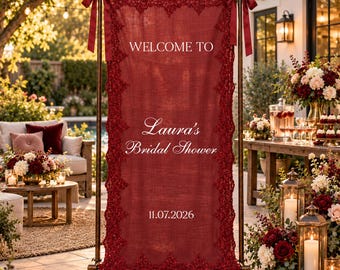 May include: A rectangular deep red fabric banner suspended from a bronze metal frame with matching ribbons. The banner features white text reading WELCOME TO Laura's Bridal Shower 11.07.2026 and is bordered by intricate dark red lace trim.