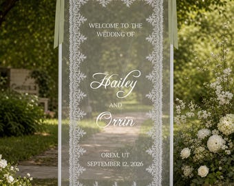 May include: A rectangular white metal frame holding a sheer white fabric panel with intricate white lace borders. The panel features the text: WELCOME TO THE WEDDING OF Hailey AND Orrin OREM, UT SEPTEMBER 12, 2026. Two sage green ribbons are tied at the top.
