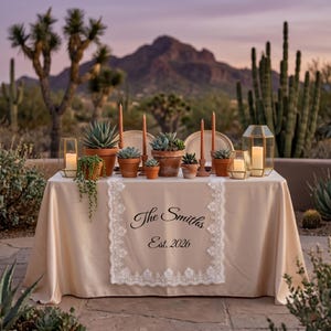 Puede incluir: Una mesa preparada para un evento al aire libre con un mantel beige y un camino de mesa de encaje blanco. El camino de mesa tiene el texto "The Smiths Est. 2026". La mesa est&aacute; decorada con suculentas en macetas, velas y faroles dorados. El fondo presenta plantas del desierto y una monta&ntilde;a.