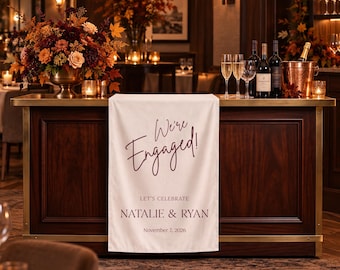 May include: A white fabric table runner draped over a dark wood bar, featuring the dark red text We're Engaged!, LET'S CELEBRATE, NATALIE & RYAN, and November 7, 2026. The runner is positioned below a large floral arrangement and wine glasses.