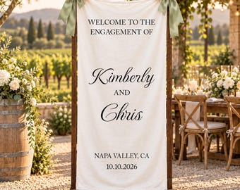 May include: A vertical off-white fabric banner suspended from a dark wooden frame, featuring sage green ribbon bows at the top corners. The text reads: WELCOME TO THE ENGAGEMENT OF Kimberly AND Chris NAPA VALLEY, CA 10.10.2026 in black lettering.