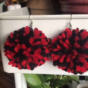 May include: A pair of red and black yarn pom-pom earrings. The earrings are round and fluffy, and they are hanging from silver hooks.