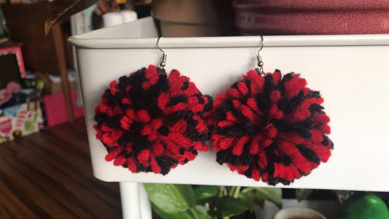 May include: A pair of red and black yarn pom-pom earrings. The earrings are round and fluffy, and they are attached to silver hooks.