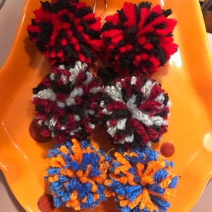 May include: Six colorful yarn pom-poms on a orange plate. The pom-poms are red and black, red and white, and orange and blue.