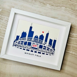 May include: A white framed print with a blue and white silhouette of Wrigley Field, a baseball stadium in Chicago, Illinois. The print includes the text "Wrigley Field" and a red heart.