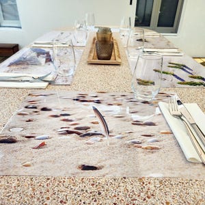 Peut inclure: Une table à manger dressée avec des sets de table représentant des scènes de plage. Chaque couvert comprend un verre à eau, des couverts et une serviette blanche. Un plateau en bois contient des objets décoratifs.