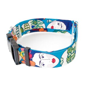 May include: A blue dog collar with a repeating pattern of stylized faces and floral designs. The collar has a black plastic buckle and a silver metal loop for attaching a leash. The collar is made of fabric.