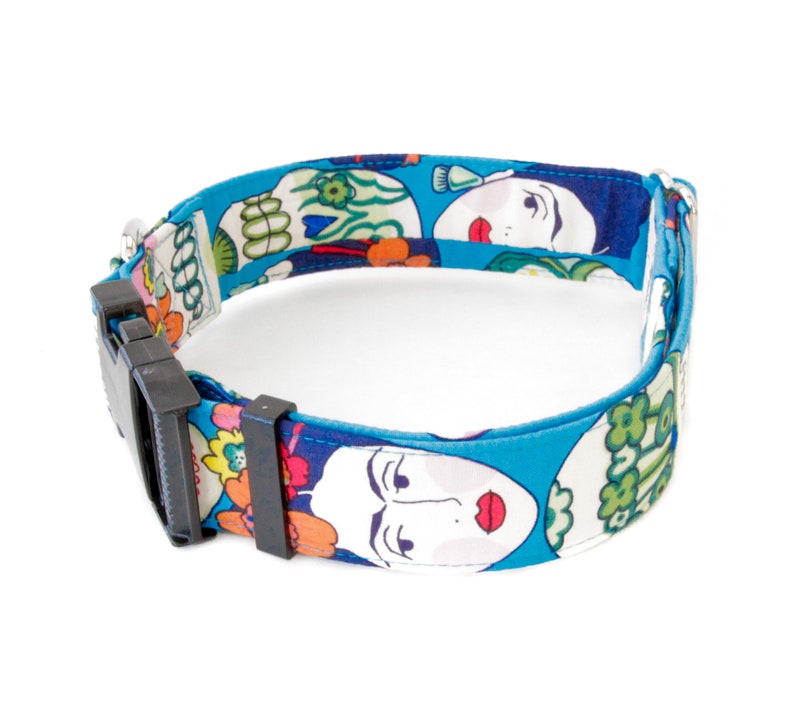 May include: A blue dog collar with a repeating pattern of stylized faces and floral designs. The collar has a black plastic buckle and a silver metal loop for attaching a leash. The collar is made of fabric.