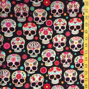 May include: Black fabric featuring a repeating pattern of colorful sugar skulls, flowers, and stars. The skulls are decorated with intricate floral designs in pink, red, yellow, and turquoise. A yellow ruler is visible on the right side.