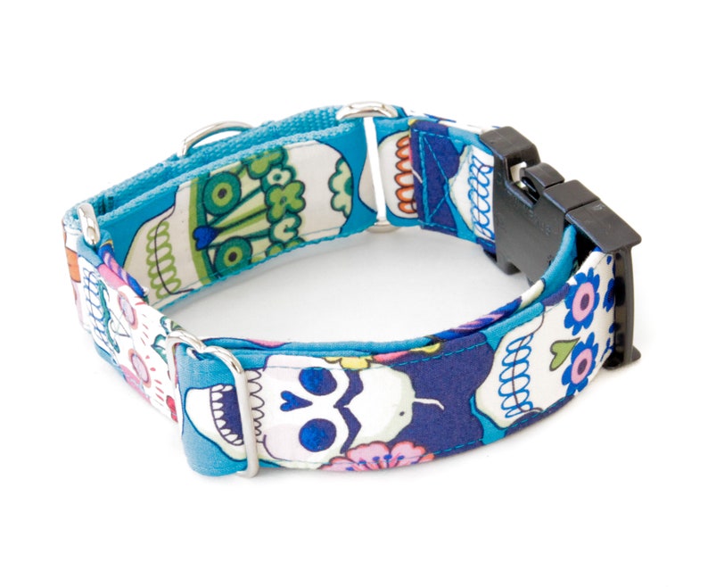 May include: A blue dog collar with a Day of the Dead skull pattern. The collar features a black plastic buckle and silver metal hardware. The fabric has a repeating pattern of skulls, flowers, and other decorative elements.