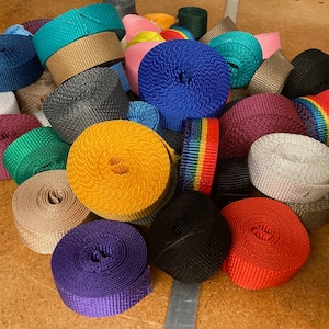 May include: A collection of colorful rolls of webbing in various colors, including blue, yellow, red, and green. The rolls are tightly wound and appear to be made of a durable, woven material. Some rolls have a rainbow stripe design. The webbing is suitable for crafting.