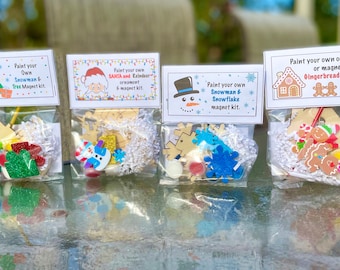 May include: Four clear plastic bags with Christmas-themed paint-your-own ornament kits. The kits include wooden ornaments, paint, glitter, and other embellishments. The bags are labeled with the names of the ornaments: Snowman & Tree, Santa and Reindeer, Snowman & Snowflake, and Gingerbread House.