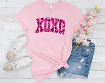 May include: Pink t-shirt with a pink glitter XOXO graphic. The shirt is laying on a white wooden surface with a pair of white sneakers, a pair of denim shorts, and a sprig of white flowers.