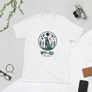 Peut inclure: T-shirt blanc avec un motif de loup hurlant à la lune dans un décor de forêt. Le motif est entouré d'un cercle avec le texte "WY*RD BRAND" en dessous.