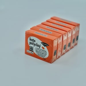 May include: A stack of six orange boxes of Kato Polyclay, an oven-hardening modeling clay. The box has a black and white label with the text "Kato Polyclay" and "Van Aken" in white. The label also includes instructions for hardening the clay in English, French, Japanese, and German.