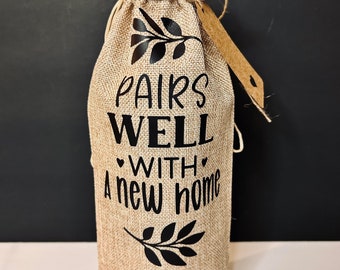 May include: A burlap wine bottle bag with black text that reads "Pairs Well With A New Home". The bag has a small house design at the bottom and a small branch design on either side of the text.