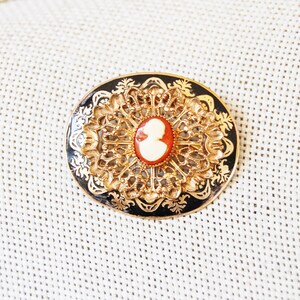 May include: A gold-toned brooch with a black enamel background and a white cameo in the centre. The cameo features a profile of a woman's face.