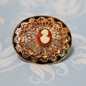 May include: A gold-toned brooch with a black enamel background and a white cameo of a woman's profile. The brooch is circular in shape and features a delicate filigree design.