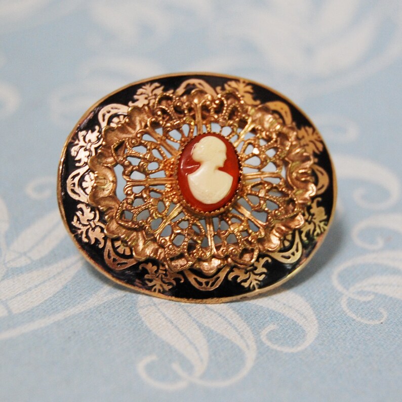 May include: A gold-toned brooch with a black enamel background and a white cameo of a woman's profile. The brooch is circular in shape and features a delicate filigree design.