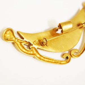 May include: A gold-toned brooch in the shape of a crescent moon with intricate details. The brooch has a pin and a safety catch on the back.