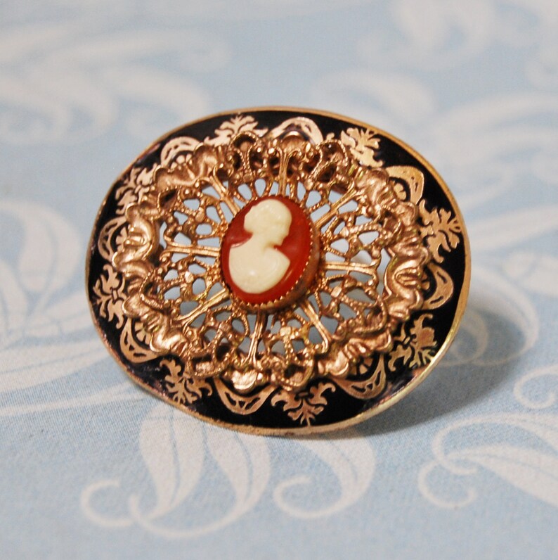 May include: A gold-toned brooch with a black enamel background and a delicate filigree design. The centre features a cameo with a white profile portrait.