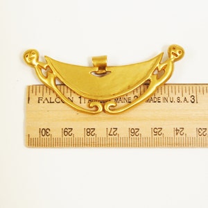 May include: Gold pendant with a crescent moon shape and two stylized figures on either side. The pendant is approximately 2.5 inches wide.
