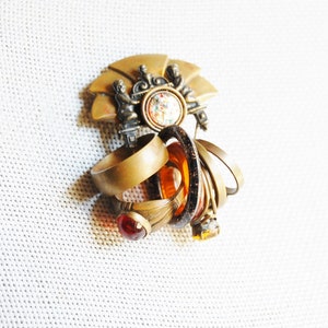 May include: A gold-toned brooch with a fan-shaped design and a central round stone. The brooch is surrounded by several rings with different colored stones, including amber and red.