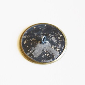 May include: A round, black button with a gold rim and a silver loop on the back.