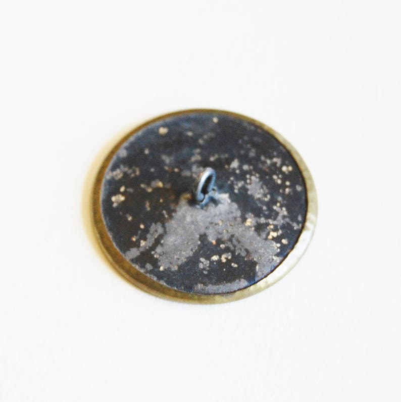May include: A round, black button with a gold rim and a silver loop on the back.