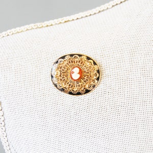 May include: A gold-toned brooch with a black enamel background and a white cameo in the centre. The cameo features a profile of a person. The brooch is oval shaped and has a delicate filigree design.