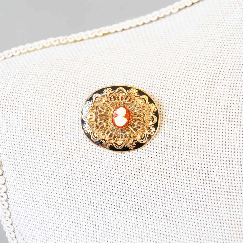 May include: A gold-toned brooch with a black enamel background and a white cameo in the centre. The cameo features a profile of a person. The brooch is oval shaped and has a delicate filigree design.