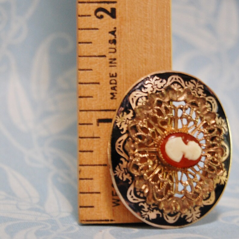 May include: A gold-toned brooch with a black and white enamel background. The centre features a cameo with a white profile image and a red background. The brooch is surrounded by a gold filigree design.