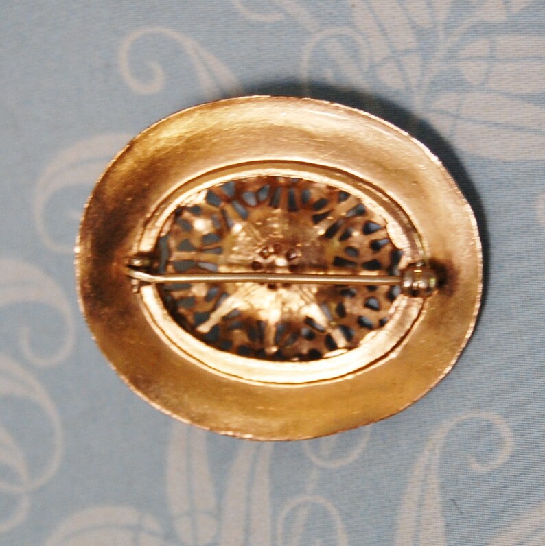 May include: A gold-toned brooch with a filigree design and a single rhinestone in the centre. The brooch has a pin and catch closure on the back.