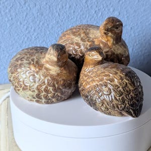 May include: Three ceramic quail figurines with brown and white speckled paint. The quail are sitting on a white circular base.