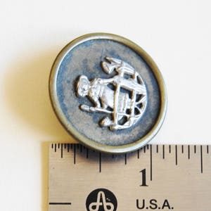 May include: A silver and brass button with a design of a person holding a spear and standing in front of a wheel. The button is approximately 1 inch in diameter.