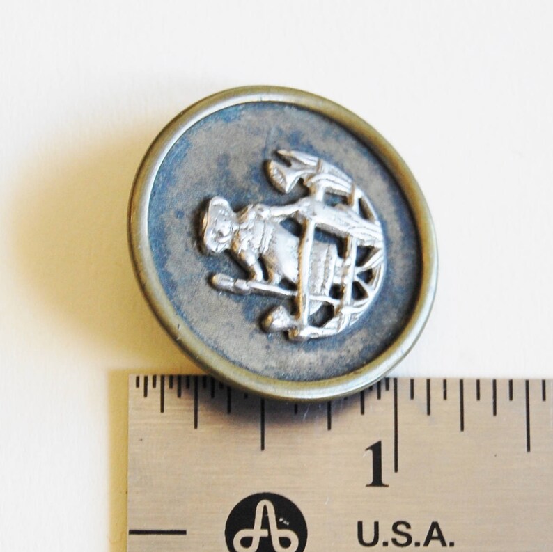 May include: A silver and brass button with a design of a person holding a spear and standing in front of a wheel. The button is approximately 1 inch in diameter.