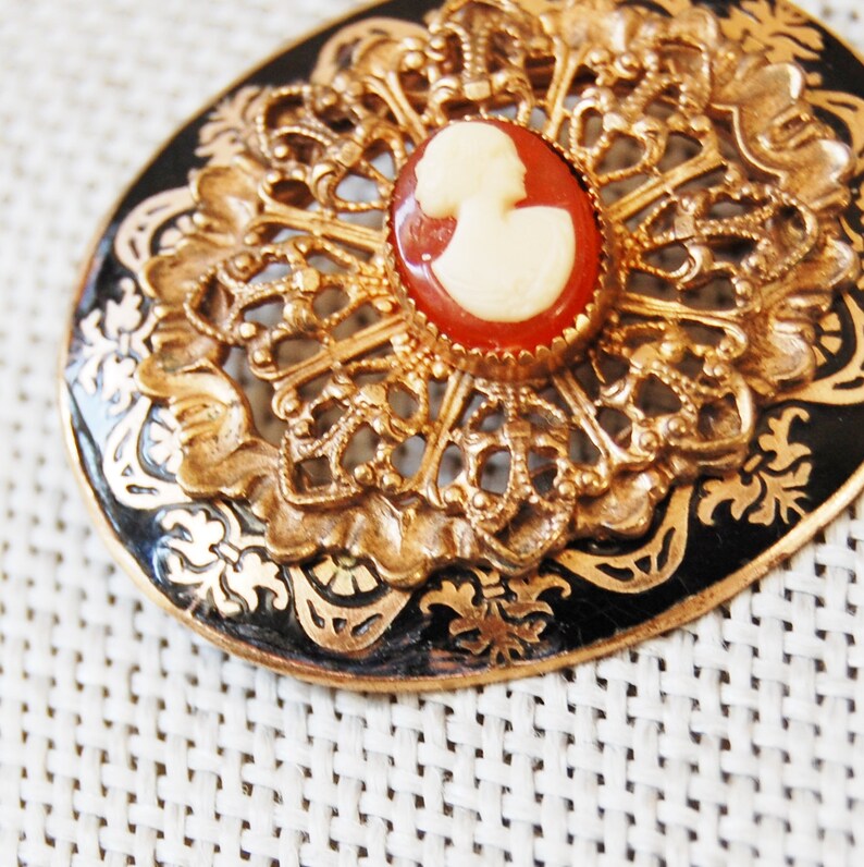 May include: A gold and black brooch featuring a cameo of a woman's profile. The cameo is set within a red and gold frame.