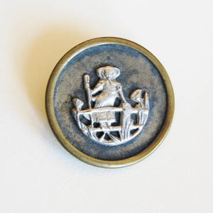 May include: A silver-toned metal button with a raised design of a woman in a dress and hat standing in a garden. The button is surrounded by a brass-colored rim.