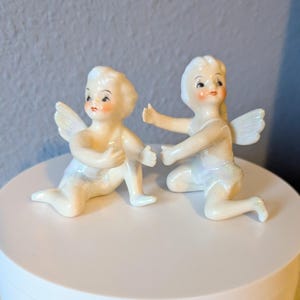 May include: Two white porcelain cherub figurines with wings. One cherub is sitting with its hands clasped together, while the other cherub is reaching out with one hand.