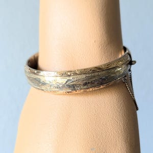 May include: A gold-toned bangle bracelet with an intricate floral design. The bracelet has a hinged clasp and a safety chain.