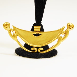 May include: A gold pendant in the shape of a crescent moon with two stylized figures on either side. The pendant has a loop at the top for hanging.
