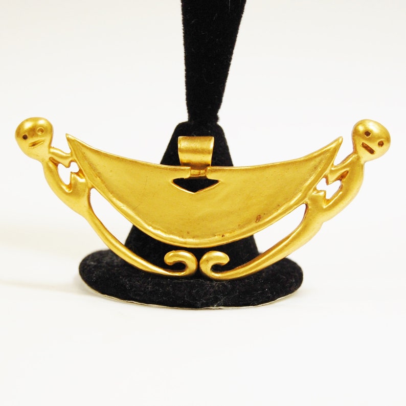May include: A gold pendant in the shape of a crescent moon with two stylized figures on either side. The pendant has a loop at the top for hanging.