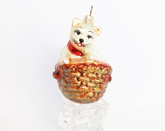 May include: A white glass Christmas ornament shaped like a cat sitting in a basket. The cat has red and white painted details and a gold glitter basket.