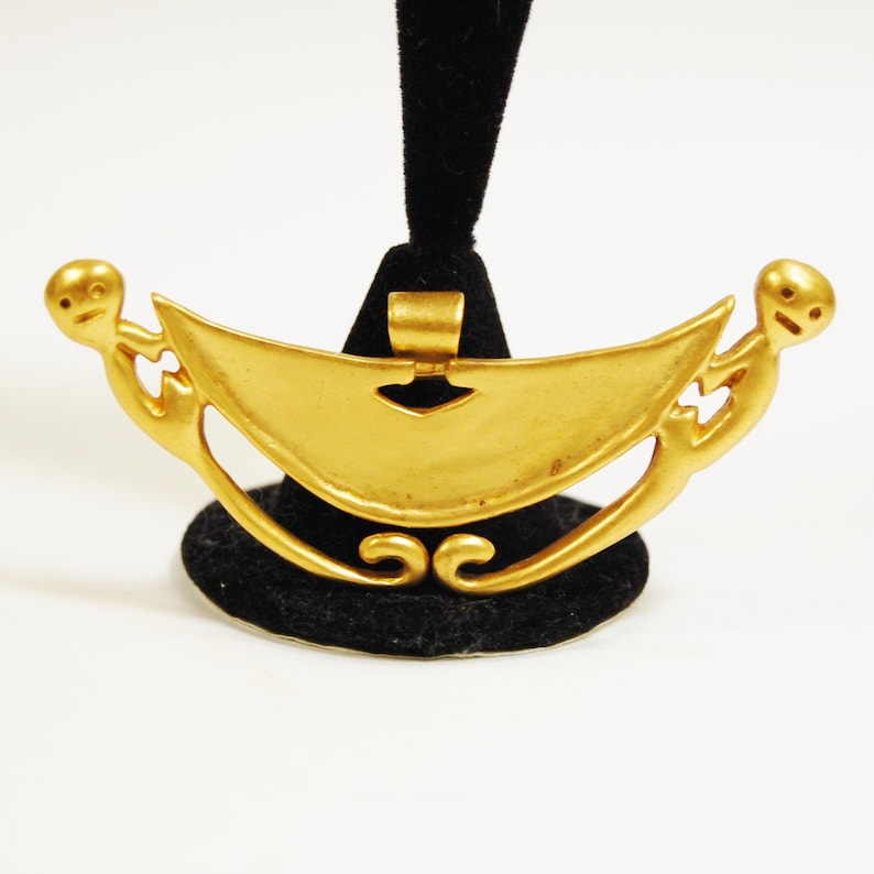 May include: Gold pendant necklace featuring two stylized figures holding a crescent moon shape. The pendant is on a black display stand.