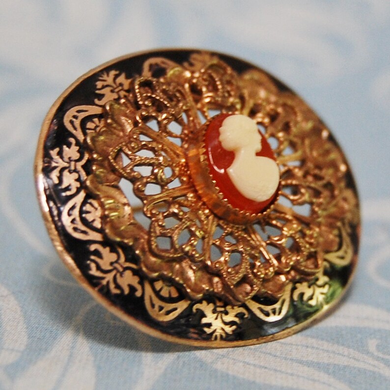 May include: A gold filigree brooch with a black enamel background and a cameo of a woman's profile in white and red.