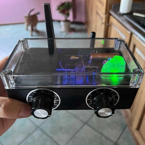 Puede incluir: Un dispositivo electrónico negro con una parte superior transparente, dos perillas y una antena. El dispositivo tiene un componente verde brillante y luces internas azules. Las perillas están numeradas. El dispositivo se sostiene en la mano de una persona.