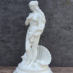 May include: A white matte sculpture of a standing figure with long flowing hair, partially covering the chest with one hand and holding a draped cloth. The figure stands on a circular base in front of a large, open seashell, rendered in solid white.
