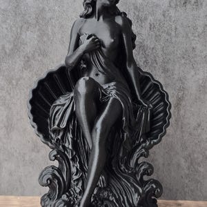 K&ouml;nnte beinhalten: Eine mattschwarze Figur, die eine Person mit langem Haar darstellt, die in einer gro&szlig;en, offenen Muschel sitzt. Die Figur ist teilweise mit Stoff bedeckt, die Beine sind gekreuzt und sie ruht auf stilisierten, wellenf&ouml;rmigen Formen auf einem runden Sockel.