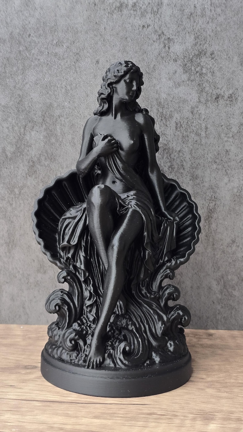K&ouml;nnte beinhalten: Eine mattschwarze Figur, die eine Person mit langem Haar darstellt, die in einer gro&szlig;en, offenen Muschel sitzt. Die Figur ist teilweise mit Stoff bedeckt, die Beine sind gekreuzt und sie ruht auf stilisierten, wellenf&ouml;rmigen Formen auf einem runden Sockel.